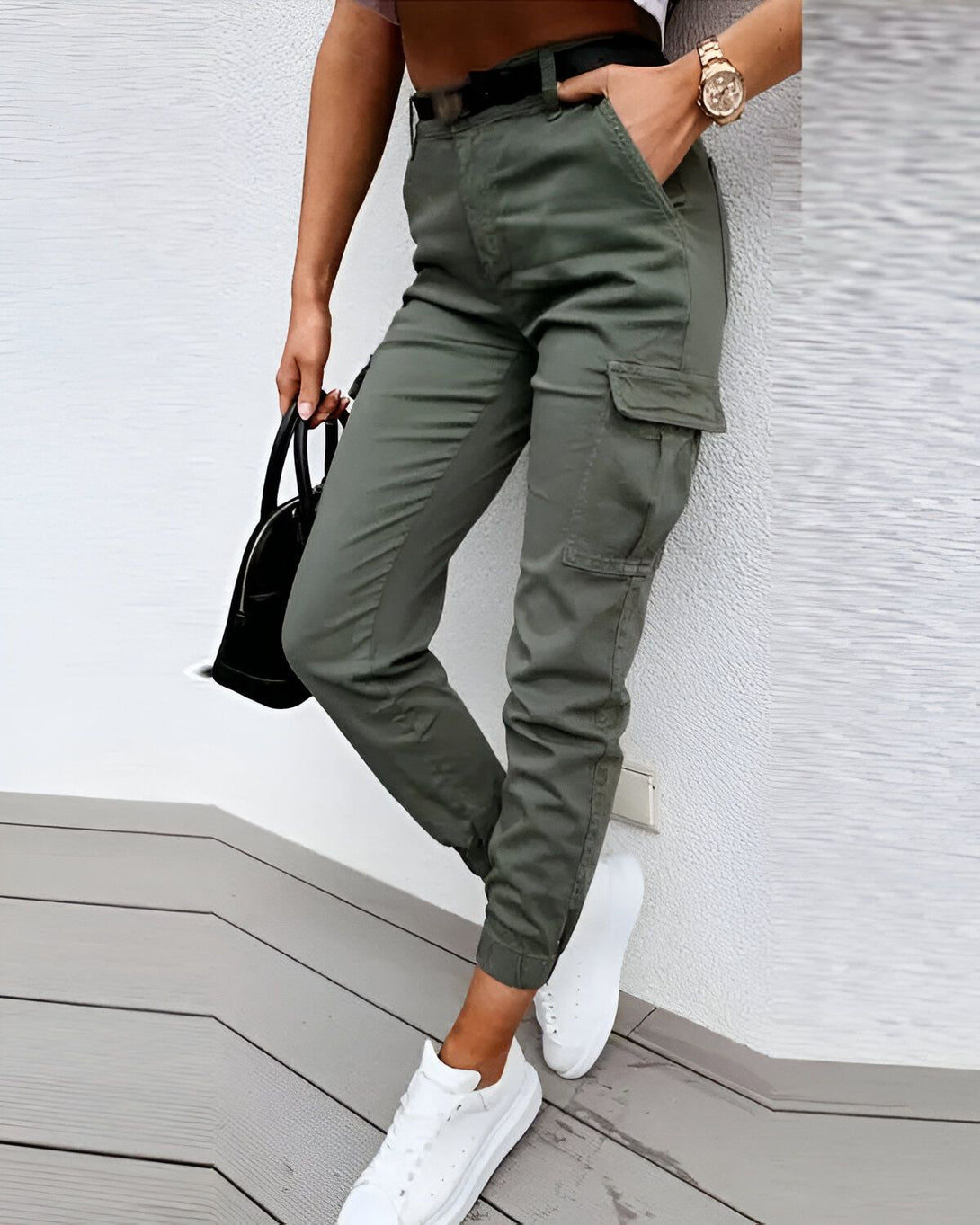 Une femme porte un pantalon cargo vert olive avec des poches cargo sur les côtés. Elle porte également un haut court noir et des baskets blanches. Elle a un sac à main noir à la main.