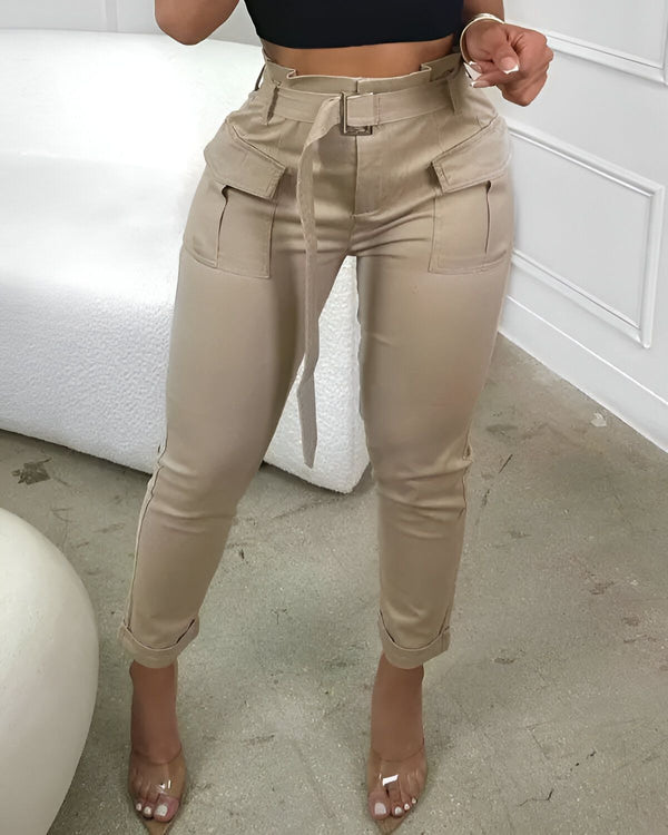 Une femme porte un pantalon cargo beige taille haute avec une ceinture. Le pantalon est ajusté aux hanches et aux cuisses, et il est légèrement évasé au niveau des chevilles. La femme porte également un haut noir et des talons transparents.