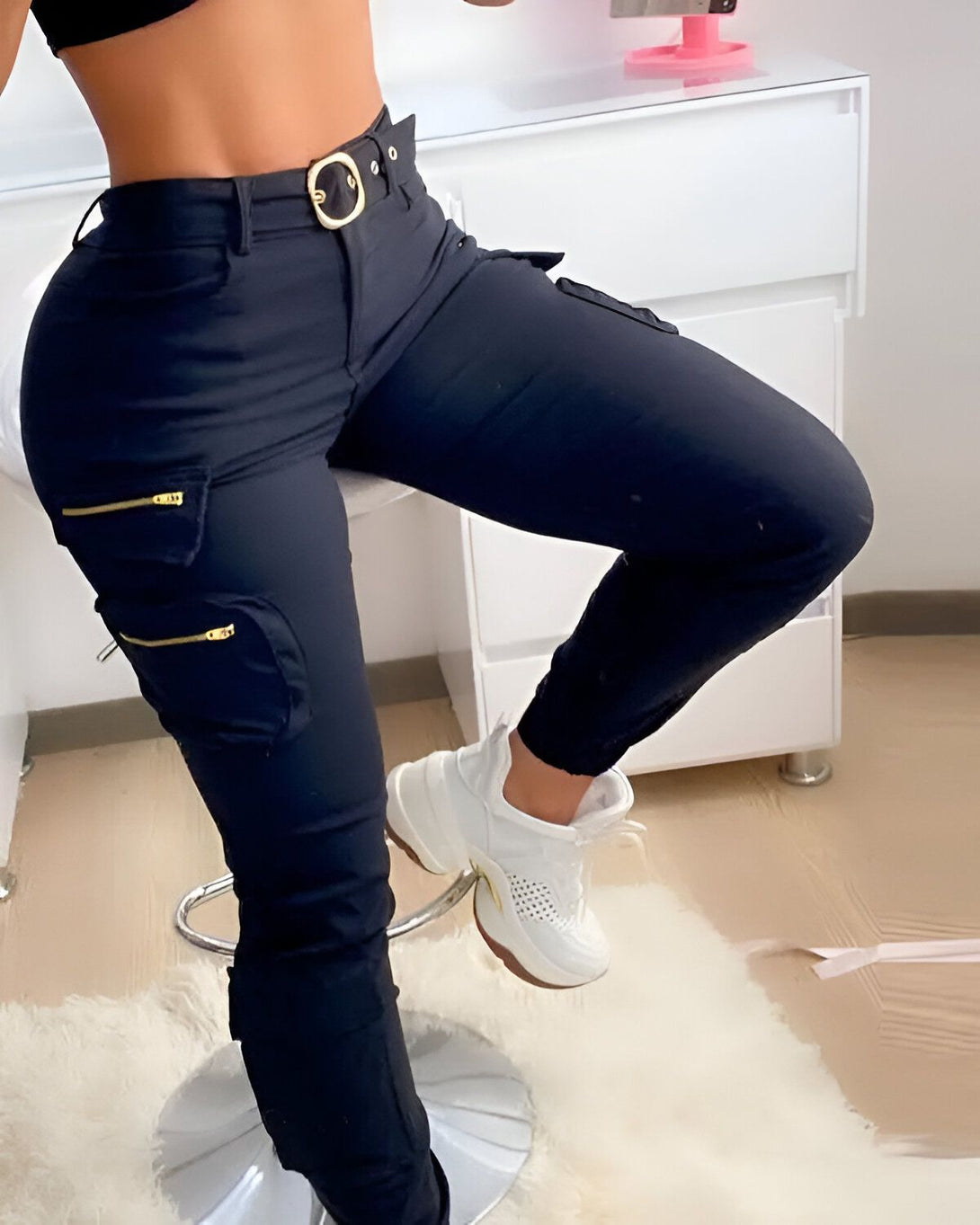 Une femme porte un pantalon cargo noir avec une ceinture dorée. Elle est debout sur un pied et l'autre jambe est posée sur une chaise. Elle porte des baskets blanches et un haut noir.