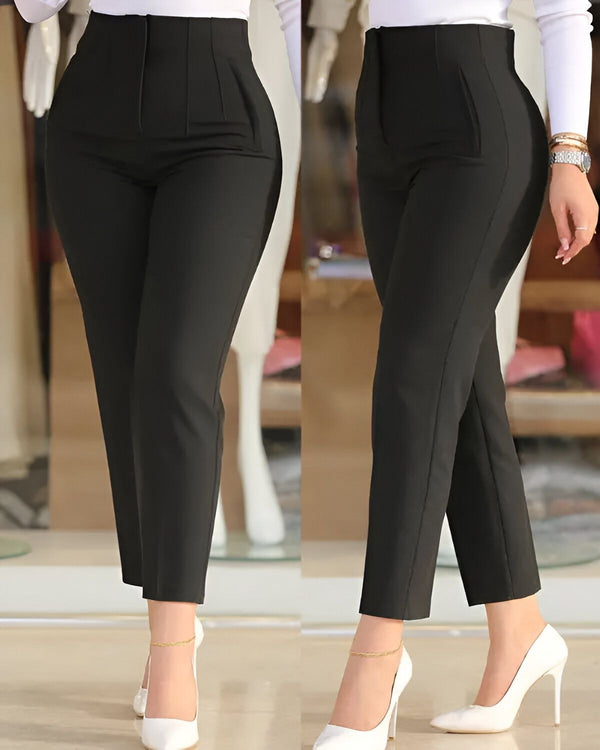 Une femme porte un pantalon noir taille haute avec des talons blancs. Le pantalon est ajusté et a une coupe droite. Il est fabriqué dans un tissu noir uni et possède deux poches latérales. La femme porte également une chemise blanche et un blazer noir. Elle a les cheveux longs et noirs et porte un maquillage léger. Elle est debout dans un magasin et sourit à la caméra.