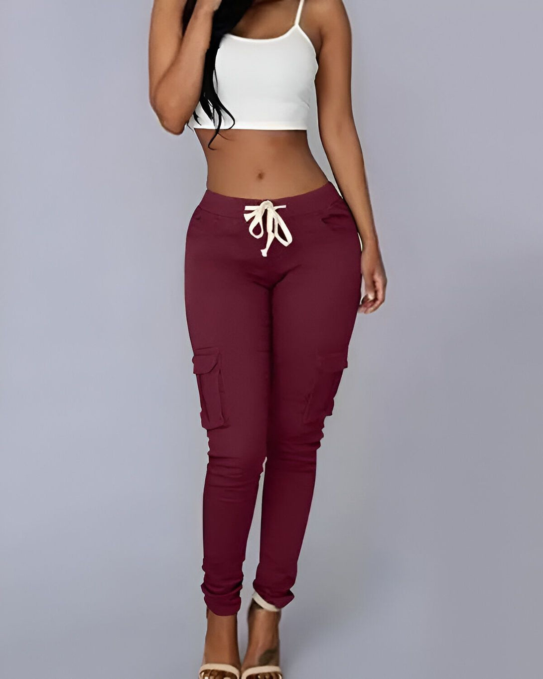 Une jeune femme porte un pantalon cargo bordeaux moulant taille haute avec des talons hauts beiges et un débardeur blanc.