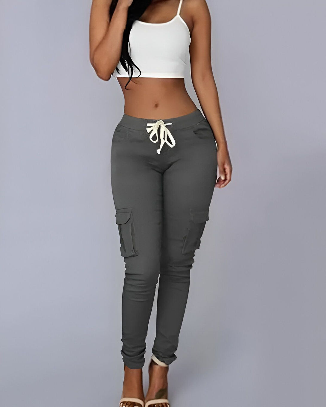 Une jeune femme porte un pantalon cargo gris taille basse avec une poche sur le côté. Elle porte également un débardeur blanc.