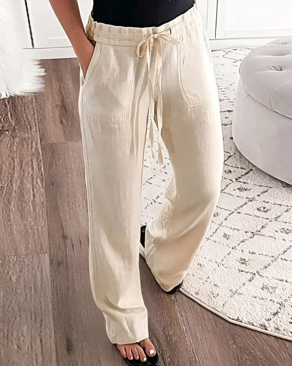 Une femme porte un pantalon large beige en lin avec une ceinture à nouer. Elle est pieds nus et se tient debout sur un tapis beige et marron. Derrière elle, il y a un pouf blanc.