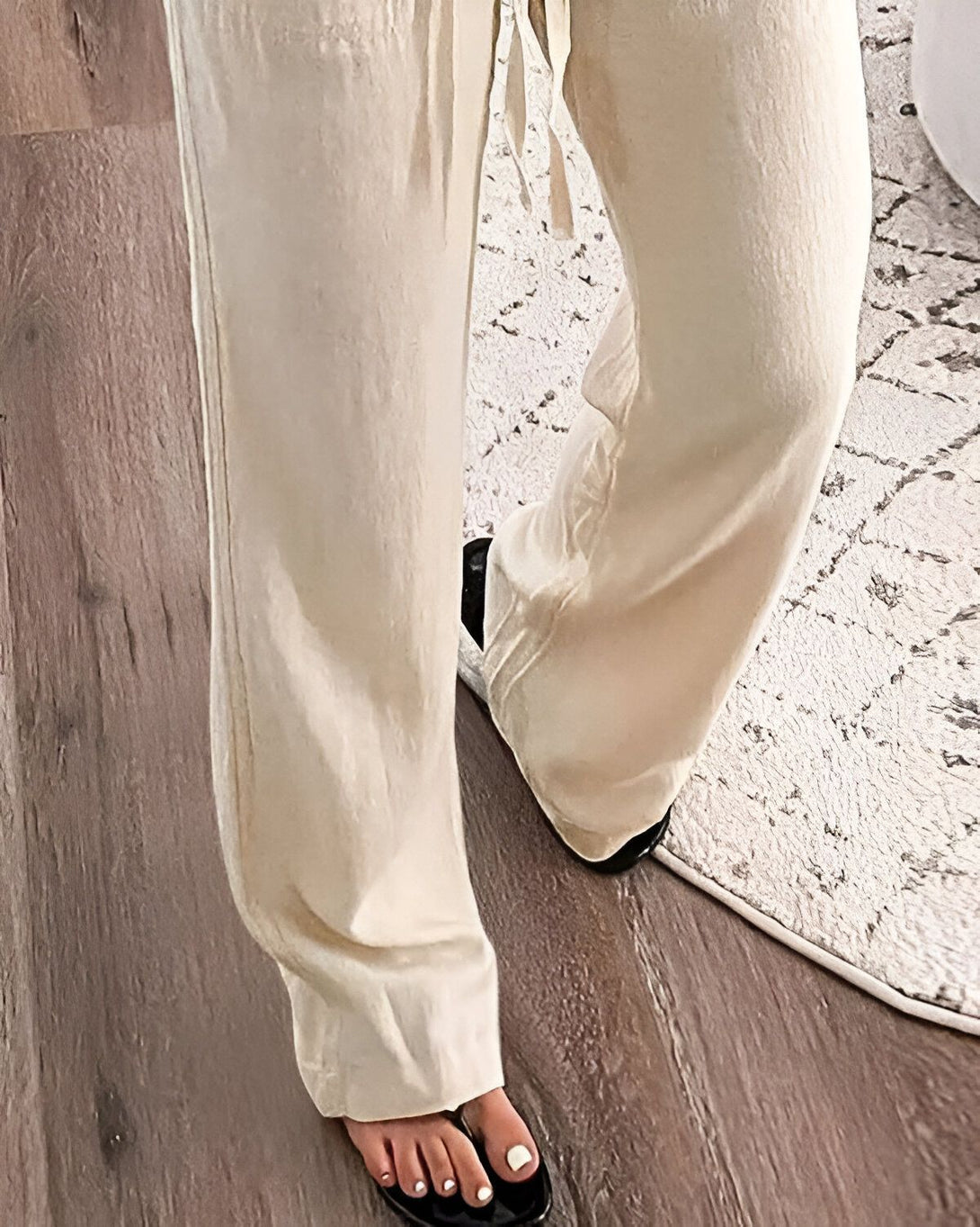 Une femme porte un pantalon beige et des sandales noires. Elle se tient debout sur un tapis à motifs noir et blanc. Le sol est en bois.