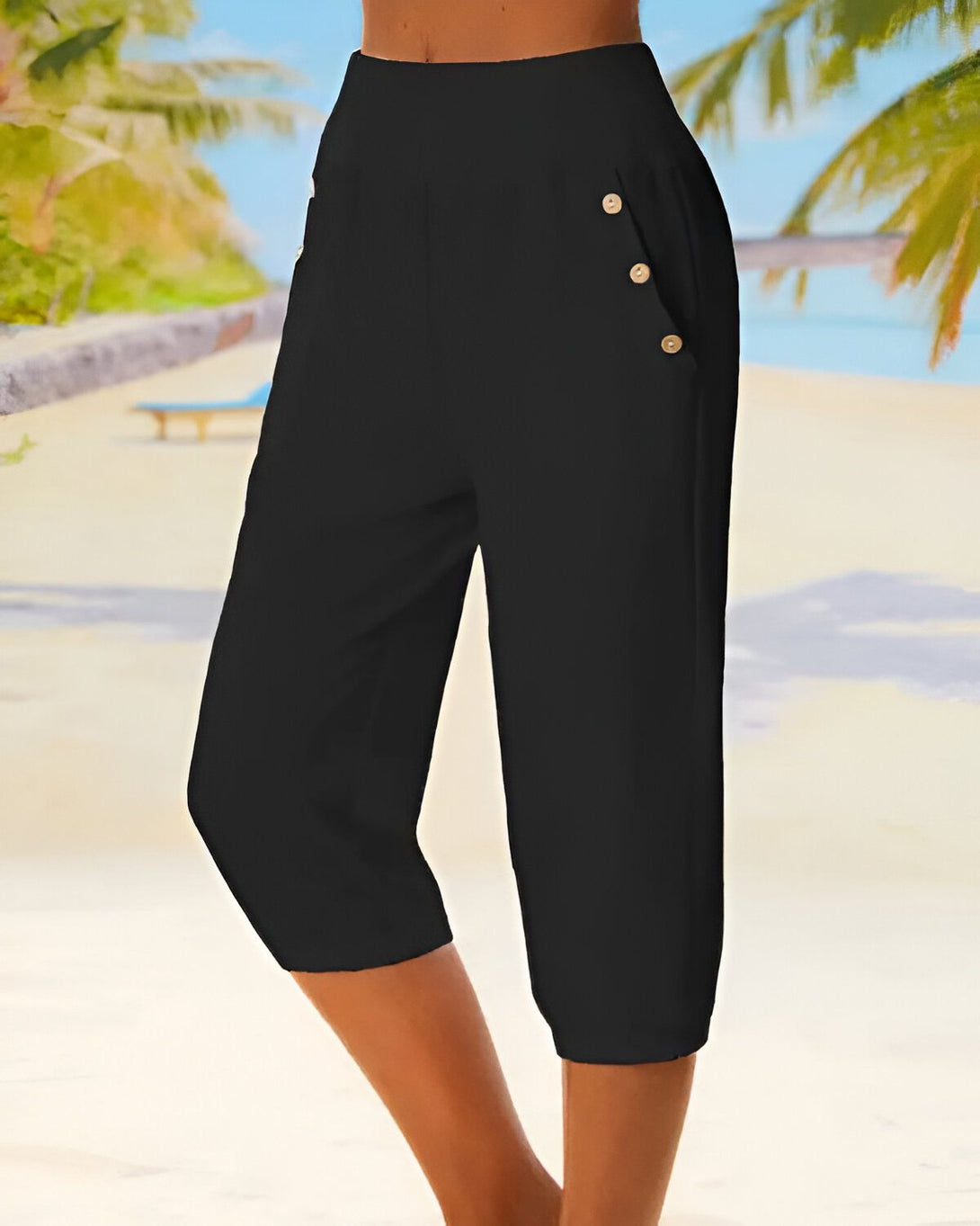 Une femme porte un pantalon noir 3/4. Le pantalon est ample et possède des boutons dorés sur le devant. La femme est debout sur une plage, dos à la mer.