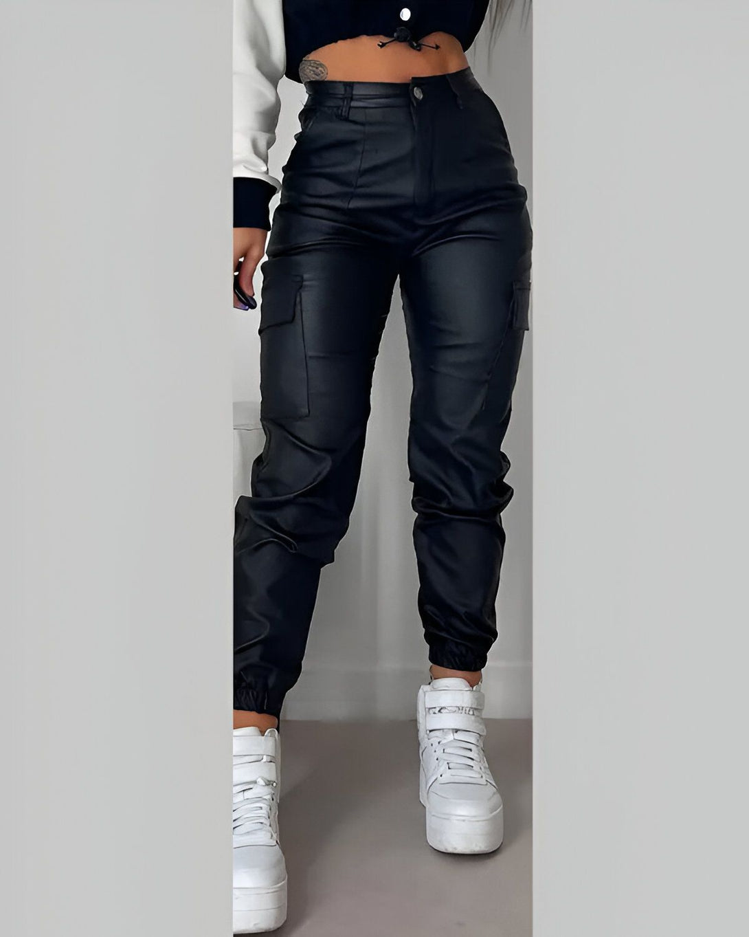 Une jeune femme porte un pantalon cargo en similicuir noir. Le pantalon est taille haute et possède des poches cargo sur les côtés. La jeune femme porte également un haut court blanc et des baskets blanches.