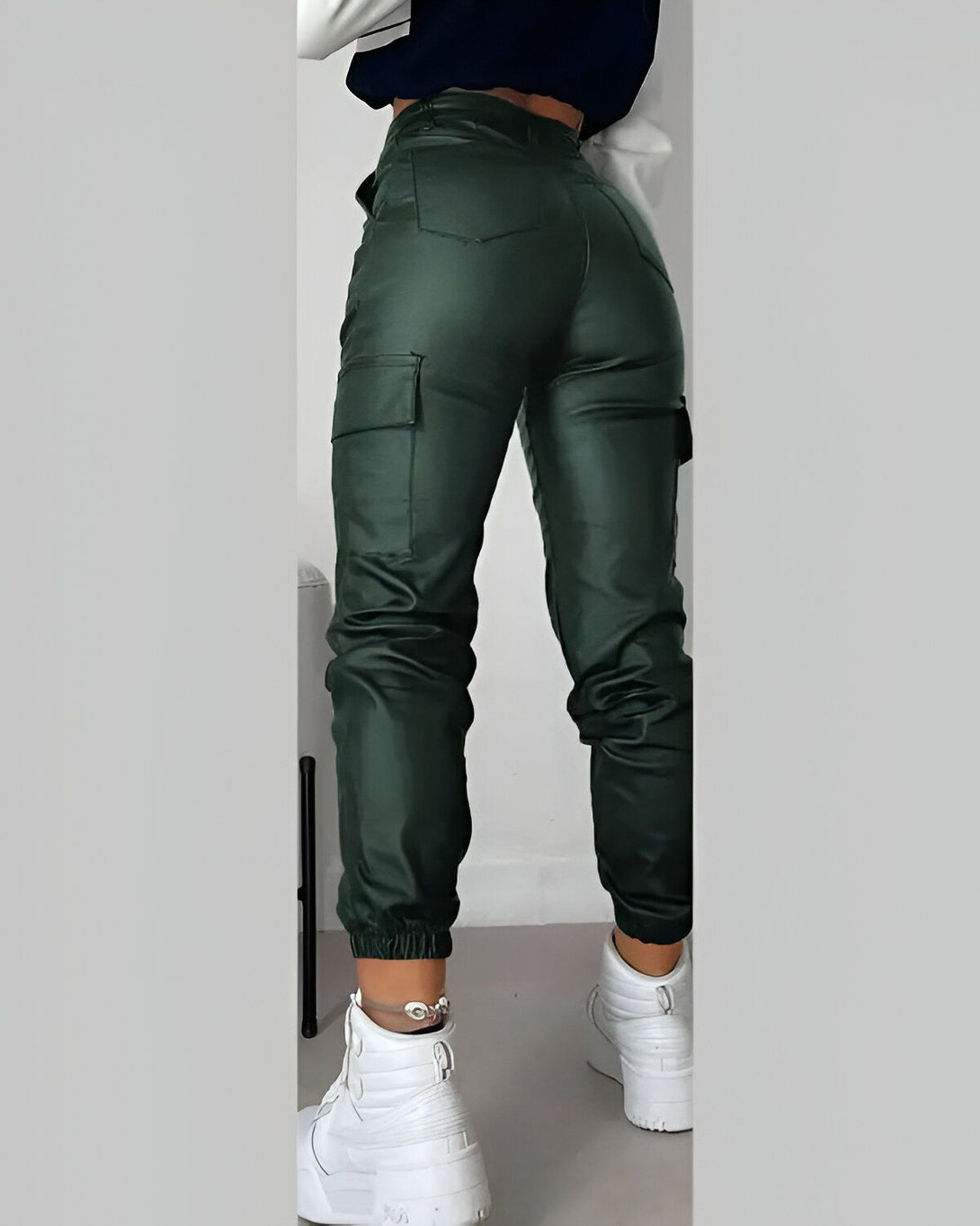 Une femme porte un pantalon cargo vert foncé en similicuir avec des poches cargo sur les côtés. Elle porte également un haut court blanc et des baskets blanches.