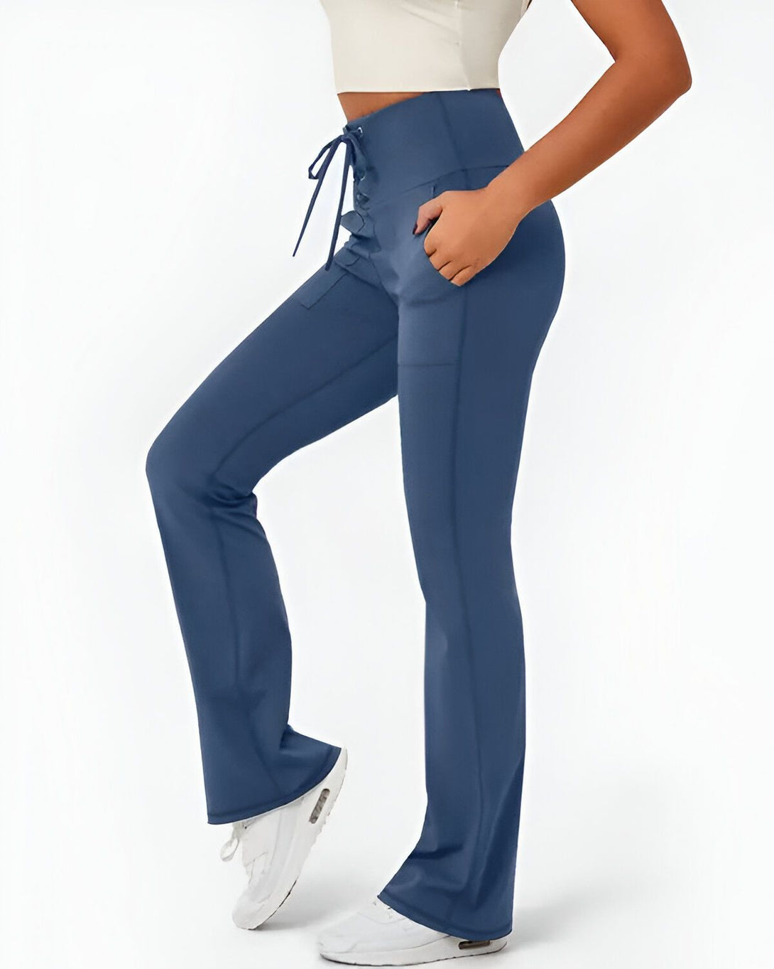 Une femme porte un legging bleu marine taille haute avec un laçage sur le devant. Le legging est évasé en bas et possède des poches sur les côtés. La femme porte également un haut blanc court et des baskets blanches.