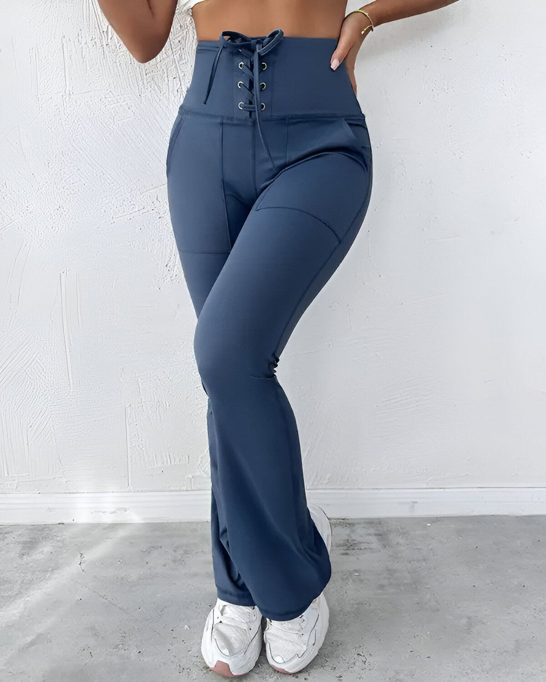 Une femme porte un pantalon de yoga bleu marine taille haute avec un laçage sur le devant. Le pantalon est évasé et possède des poches. Elle porte également un haut court blanc et des baskets blanches.