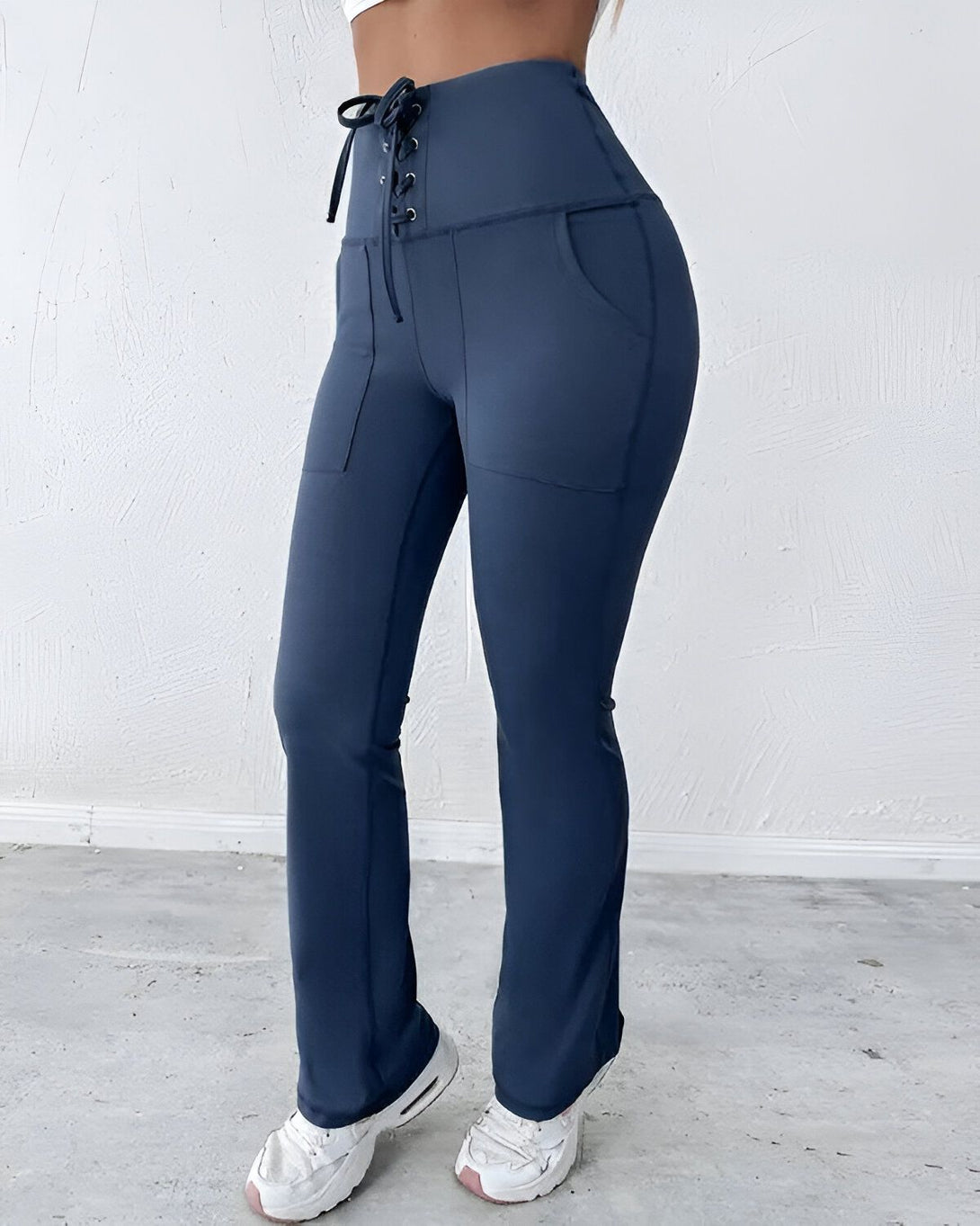 Une femme porte un legging bleu marine taille haute avec un laçage à la taille. Le legging est évasé en bas et possède des poches sur les côtés. La femme porte également des baskets blanches.
