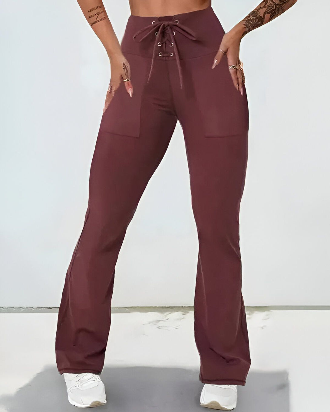 Une femme porte un pantalon marron taille haute avec un laçage sur le devant. Le pantalon est évasé et possède des poches sur les côtés. Elle porte également un haut court marron et des baskets blanches.