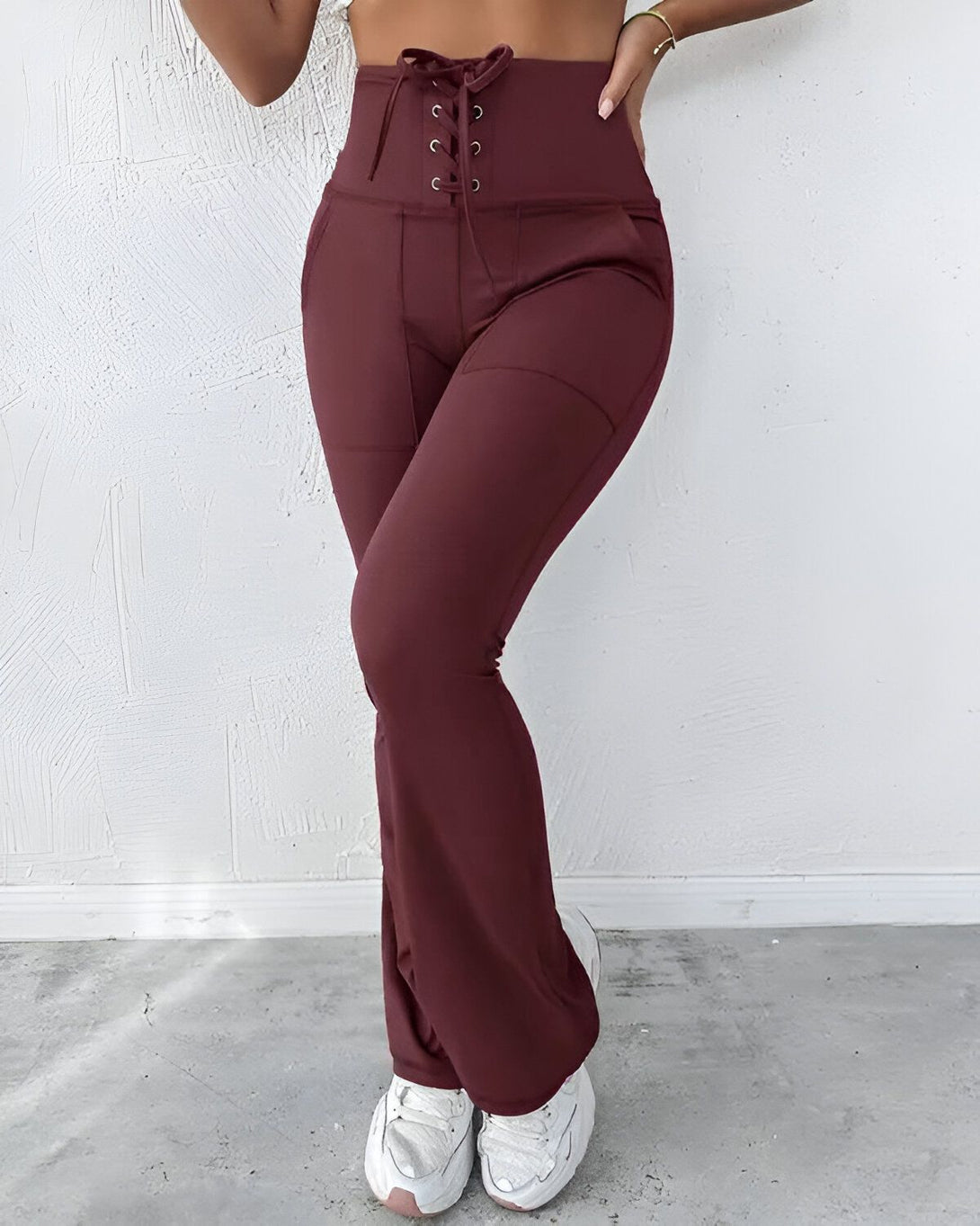 Une femme porte un pantalon marron taille haute avec un laçage sur le devant. Le pantalon est évasé et possède des poches sur les côtés. Elle porte également des baskets blanches et un haut court marron.