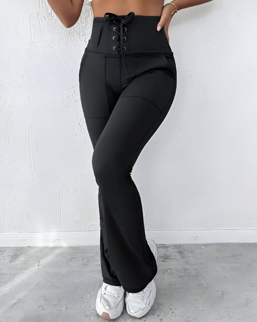 Une femme porte un pantalon noir taille haute avec un laçage à la taille. Le pantalon est évasé et possède des poches sur les côtés. La femme porte également un haut court noir et des baskets blanches.