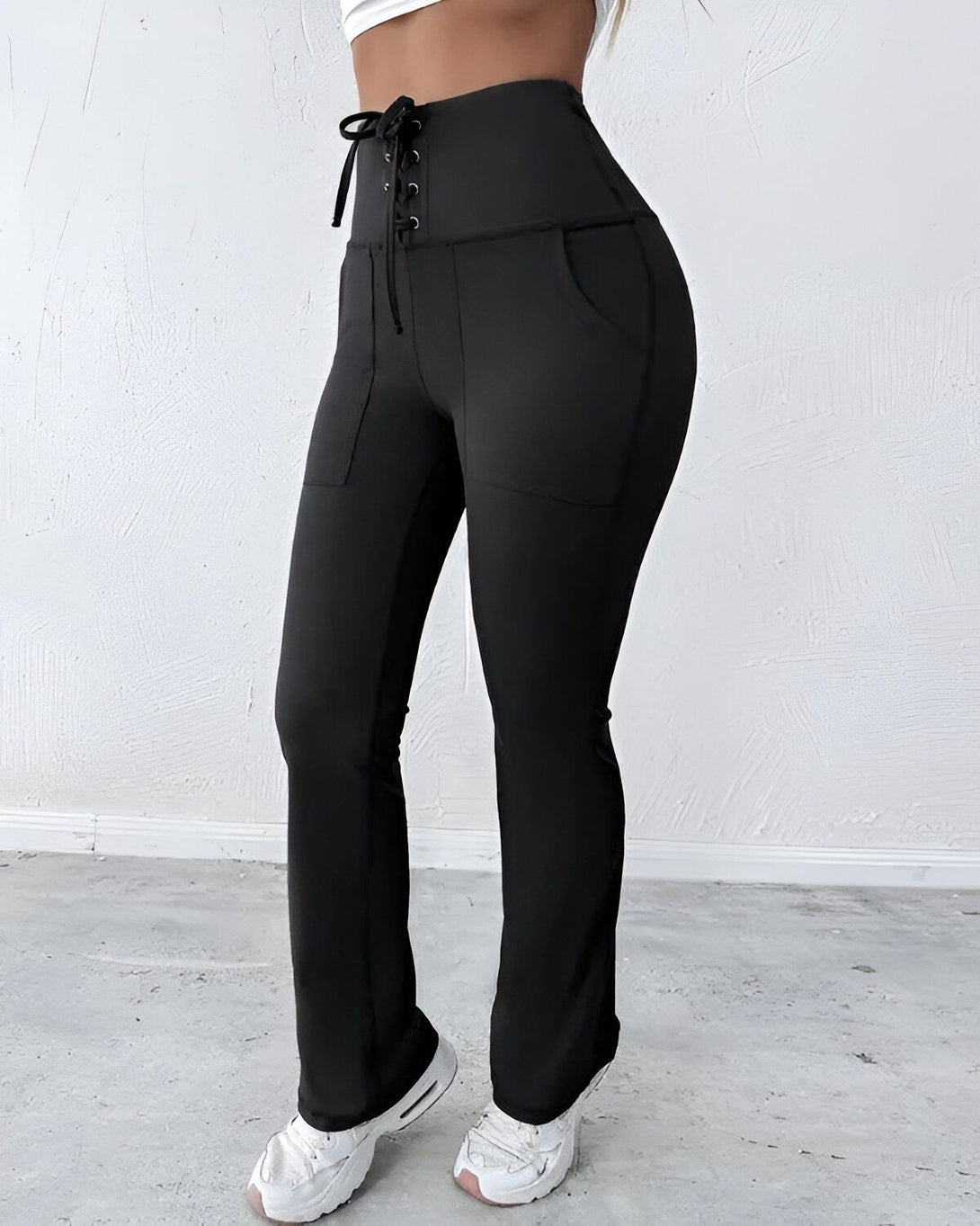 Une femme porte un pantalon de yoga noir avec un laçage à la taille. Le pantalon est taille haute et évasé. Elle porte également un haut blanc et des baskets blanches.