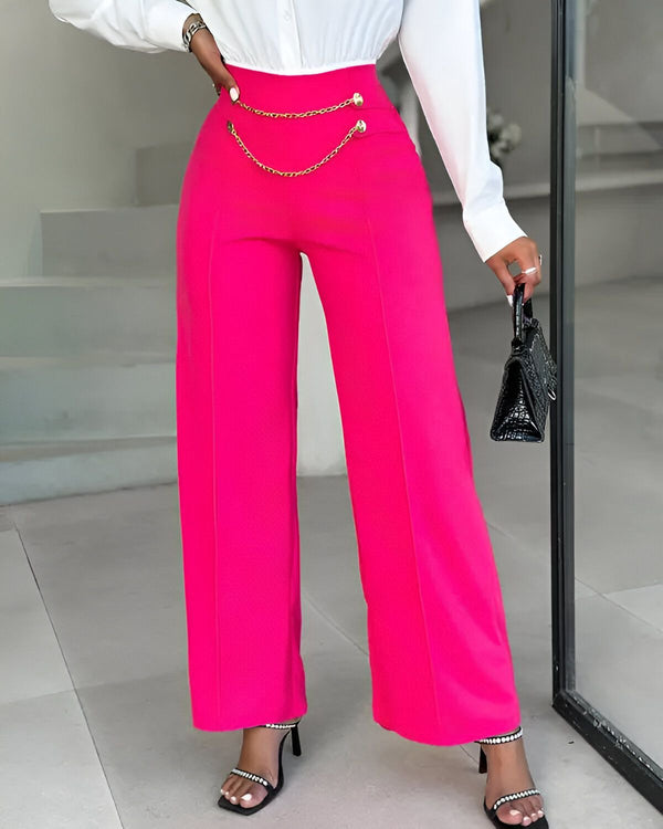Une femme porte un pantalon rose taille haute avec une chaîne dorée sur le devant. Elle porte également un chemisier blanc et des sandales noires.