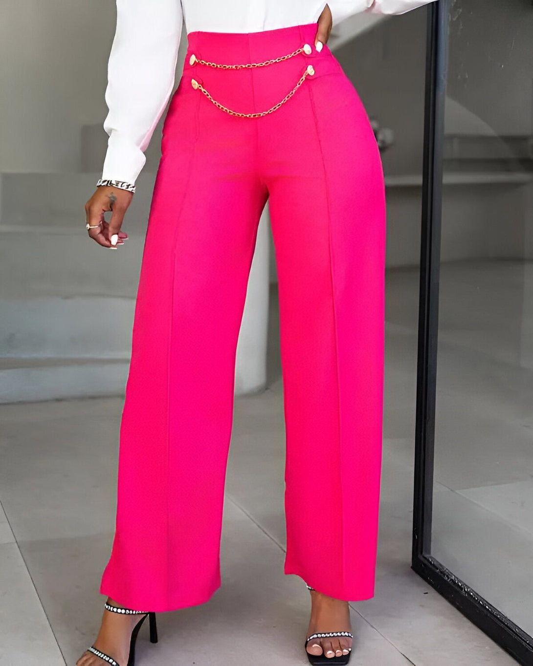 Une femme porte un pantalon rose taille haute avec une chaîne dorée à la taille. Elle porte également un chemisier blanc et des sandales à talons noirs.
