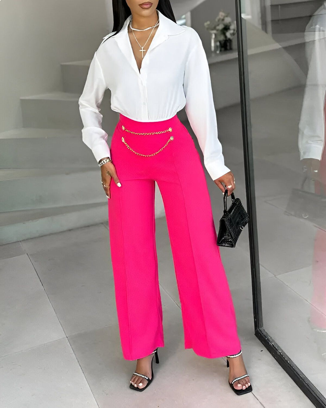 Une femme porte un pantalon rose fuchsia taille haute et une chemise blanche ouverte. Elle porte également des sandales à talons noires et un sac à main noir.