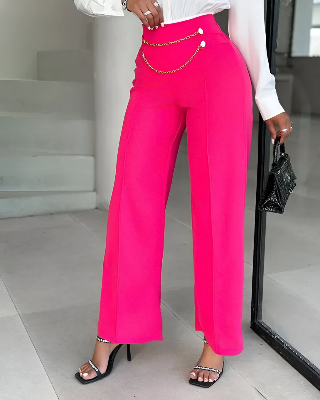 Une femme porte un pantalon large rose fuchsia taille haute avec une chaîne dorée à la taille. Elle porte également un chemisier blanc et des sandales à talons noires.