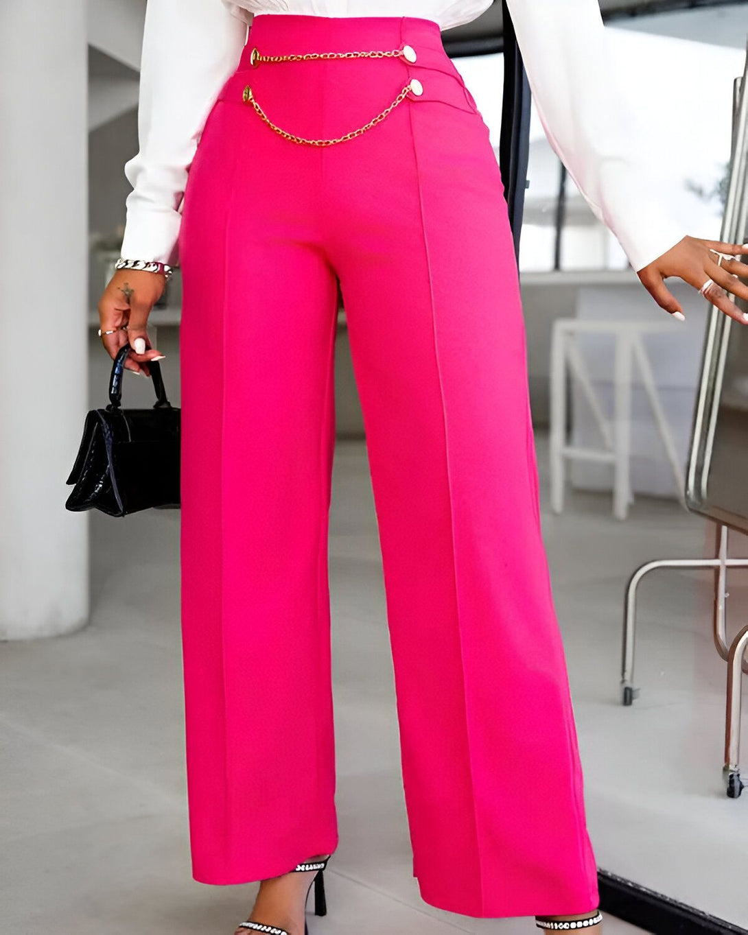 Une femme porte un pantalon rose taille haute avec une chaîne dorée à la taille. Elle porte également un chemisier blanc et des sandales à talons hauts noires.