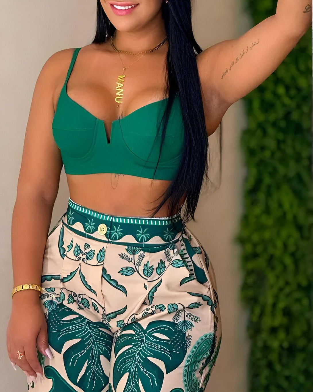 Une femme porte un haut vert et un pantalon beige à motifs de feuilles tropicales. Elle a les cheveux longs et noirs et porte un collier doré.