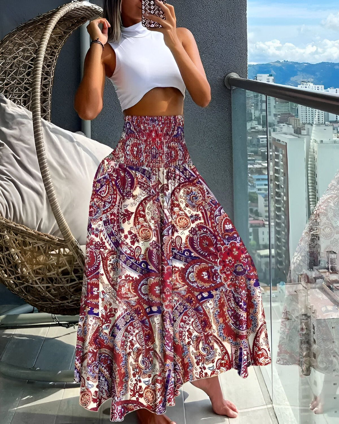 Une femme porte une longue jupe taille haute à motifs cachemire. Elle est associée à un crop top blanc. La femme est pieds nus et se trouve sur un balcon.
