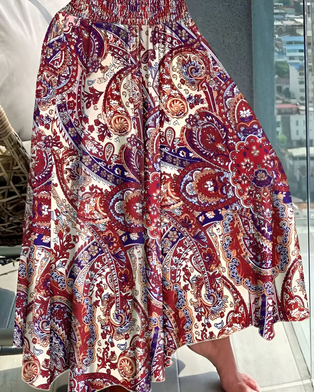 Une femme porte une jupe longue rouge à motifs cachemire. La jupe est ample et évasée, et elle est dotée d'une taille élastiquée. La femme est pieds nus et se tient sur un balcon.