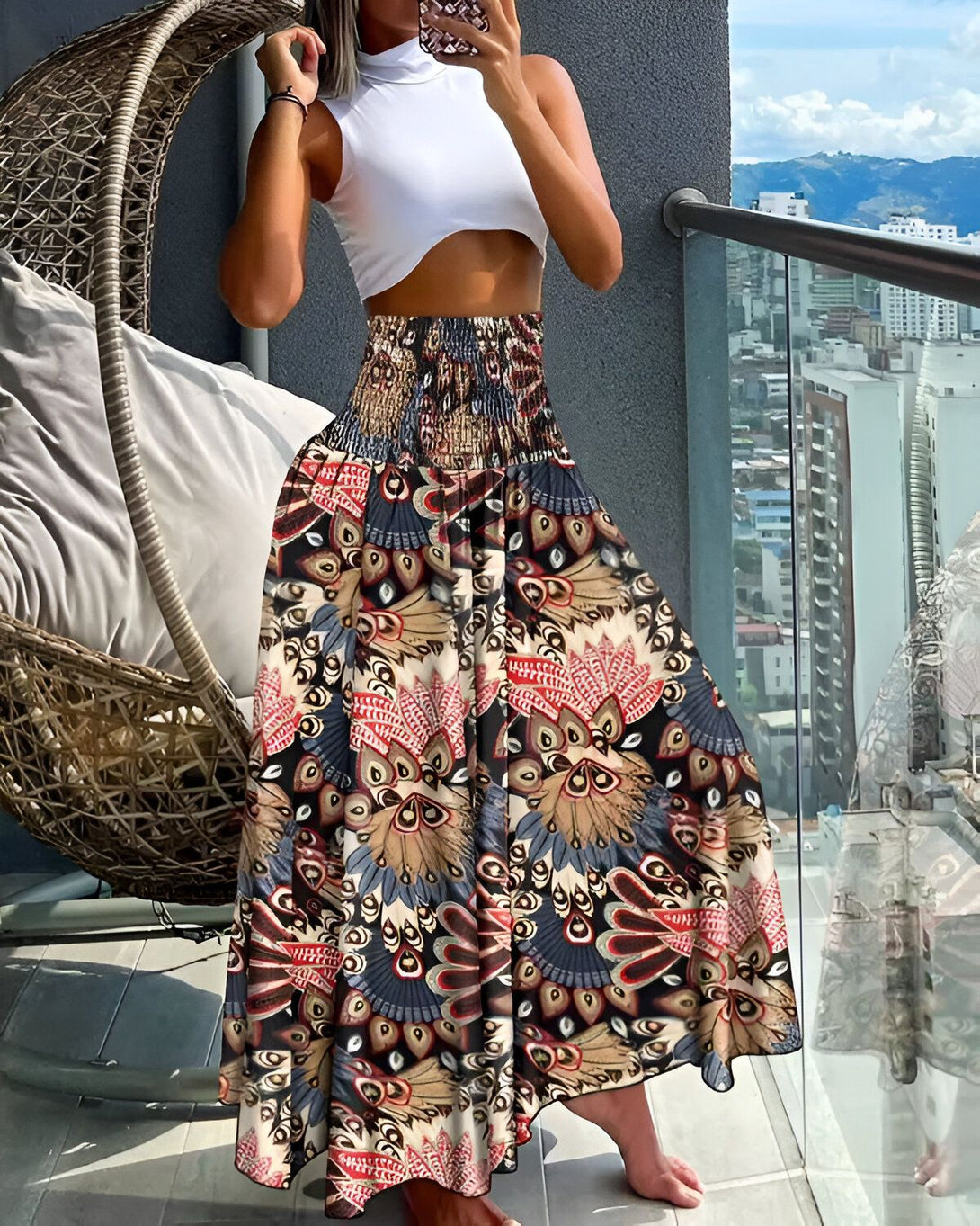 Une femme porte une jupe longue et ample à taille haute. La jupe est marron et présente un motif floral coloré. La femme est pieds nus et porte un haut blanc court. Elle se tient sur un balcon et regarde la vue.