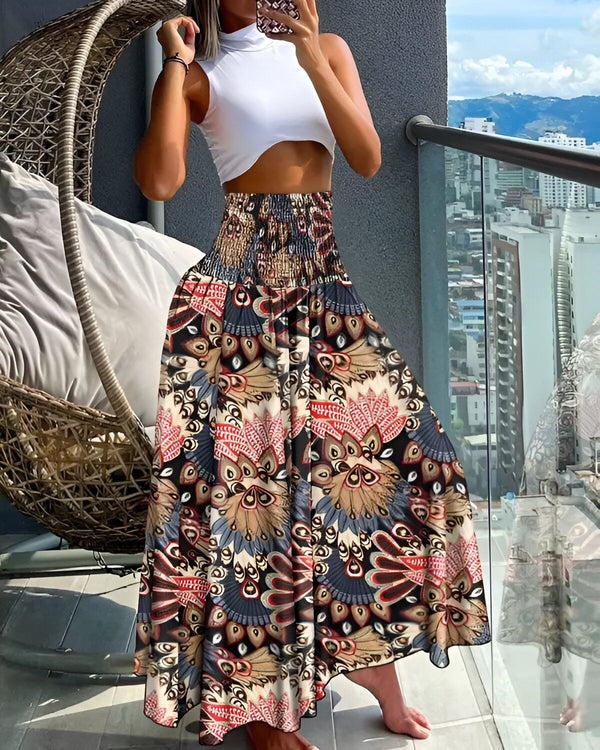 Une femme porte une jupe longue et ample à taille haute. La jupe est marron et présente un motif floral coloré. La femme est pieds nus et porte un haut blanc court. Elle se tient sur un balcon et regarde la vue.