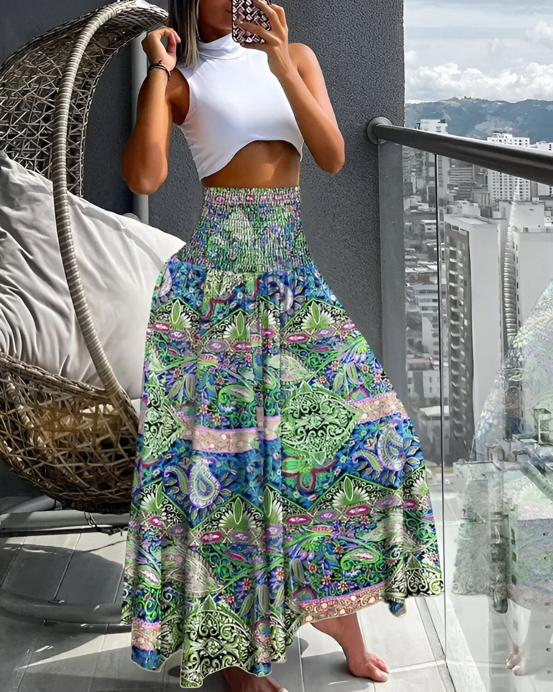Une femme porte une longue jupe colorée à motifs floraux et un haut blanc. Elle est pieds nus et se tient sur un balcon avec des montagnes en arrière-plan.