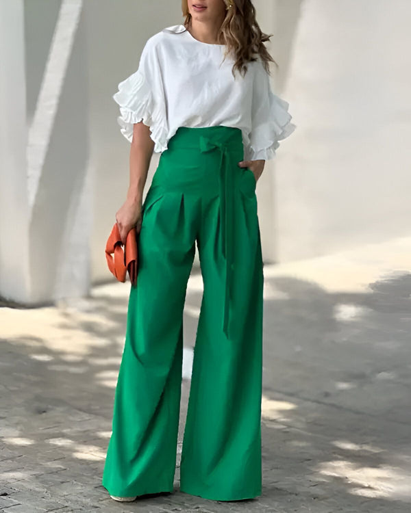 Une femme porte un chemisier blanc à manches courtes et à volants avec un pantalon vert taille haute et large. Elle porte également une ceinture marron et tient un sac à main orange.