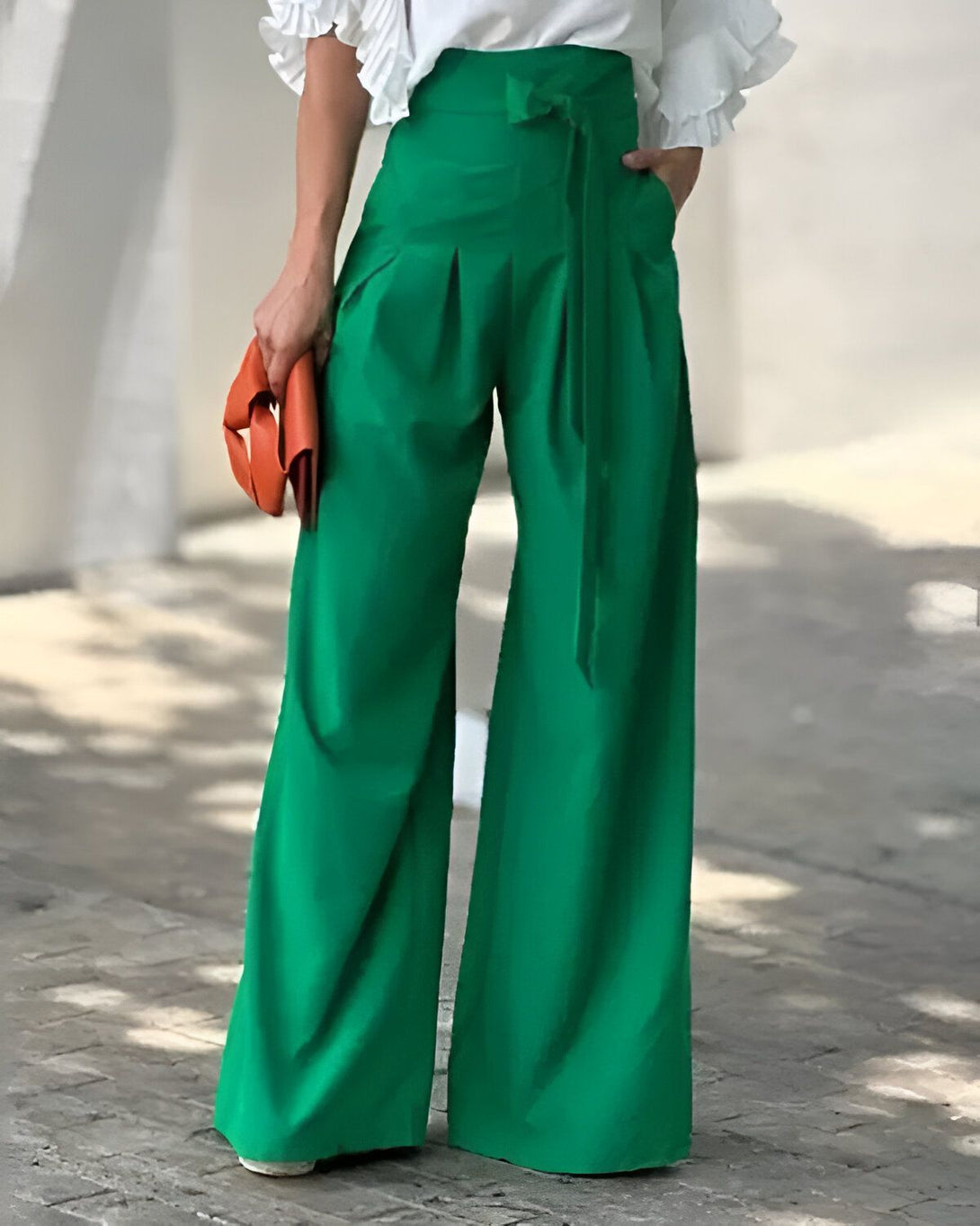 Une femme porte un pantalon large vert taille haute avec une ceinture à la taille. Elle porte également un chemisier blanc à manches bouffantes et des escarpins blancs. Elle a un sac à main orange à la main.