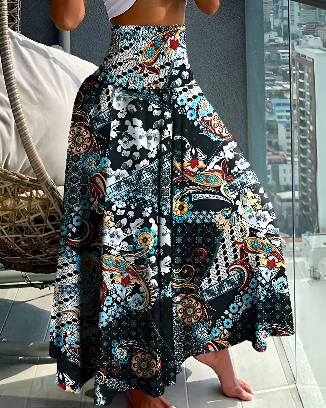 Une femme porte une jupe longue et ample à taille haute. La jupe est noire avec des motifs colorés. La femme est debout sur un balcon et regarde la vue.