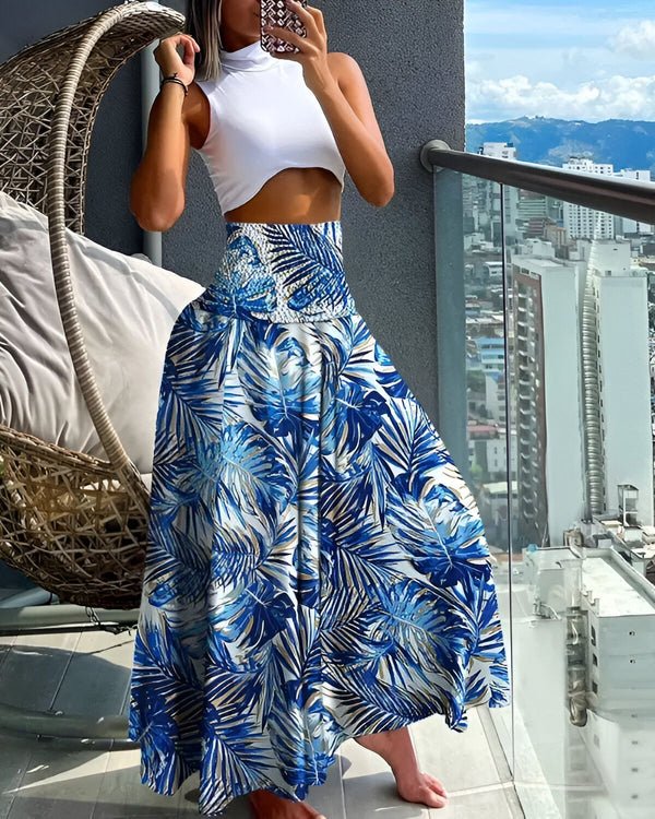 Une femme porte un haut blanc et une jupe longue bleue à motifs de feuilles tropicales. Elle est debout sur un balcon et tient son téléphone portable à la main.