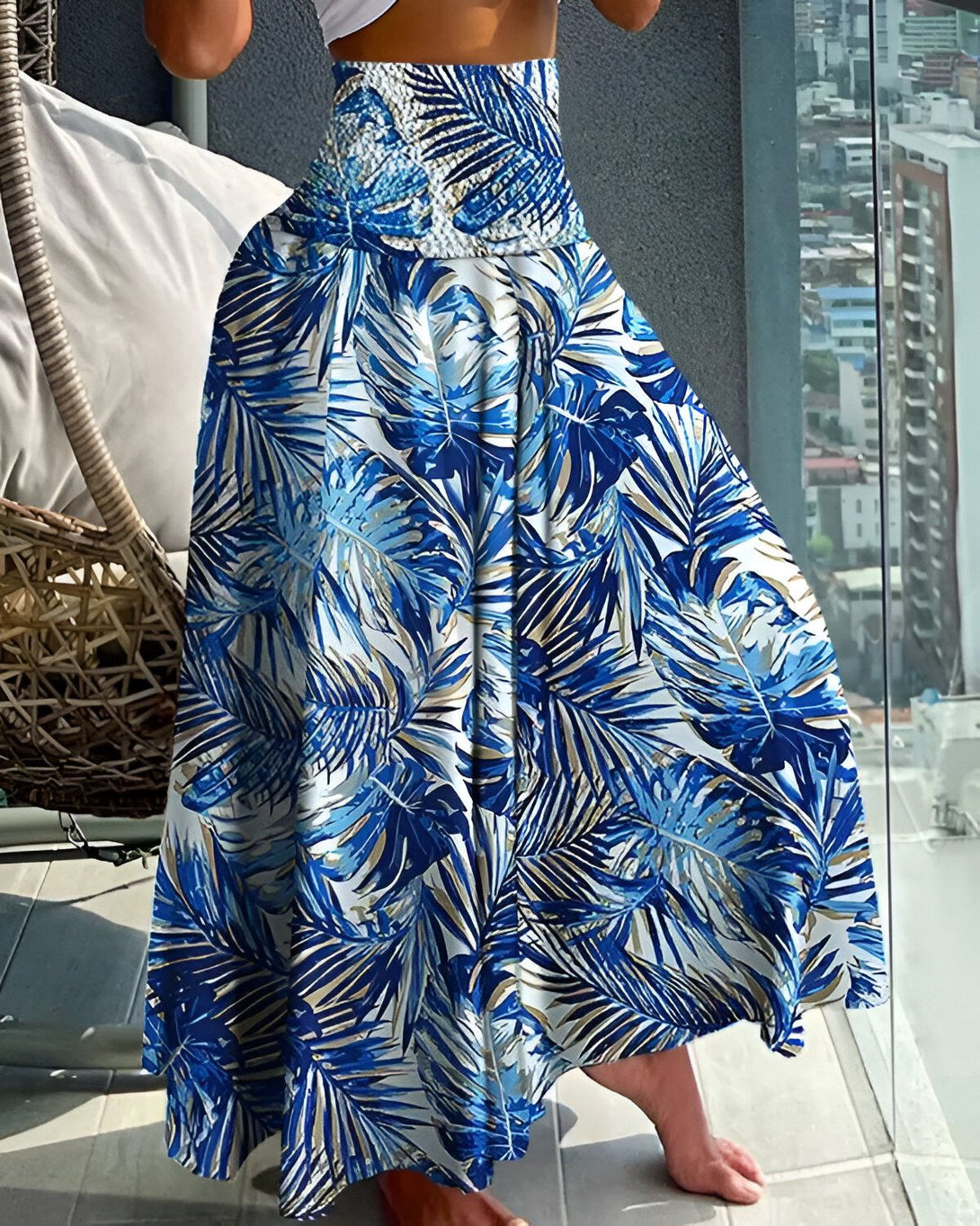 Une femme porte une jupe longue ample à taille haute. La jupe est bleue avec un motif de feuilles tropicales blanches et bleues. La femme est pieds nus et se tient sur un balcon avec une vue sur la ville en arrière-plan.