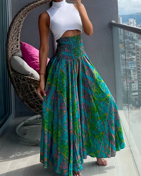 Une femme porte une longue jupe verte à motifs floraux et un haut blanc. Elle est debout sur un balcon et tient un sac à main rose.