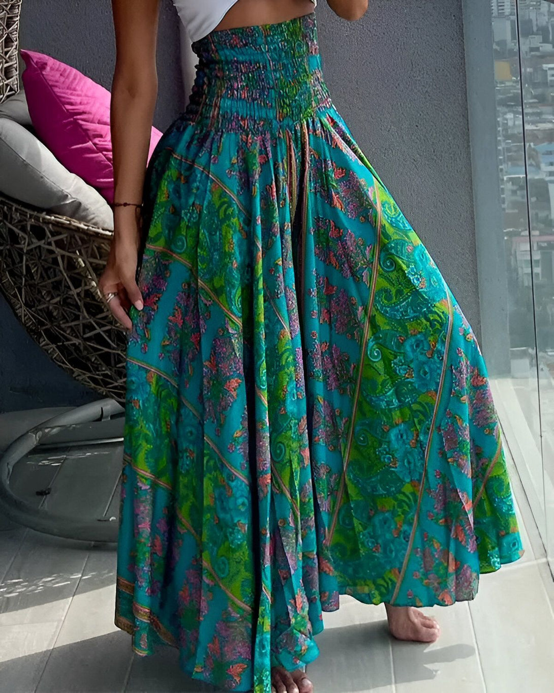 Une femme porte une longue jupe ample à taille haute. La jupe est verte avec des motifs fleuris colorés. La femme est pieds nus et porte un haut blanc. Elle se tient debout sur un balcon et l'on peut voir la ville en arrière-plan.
