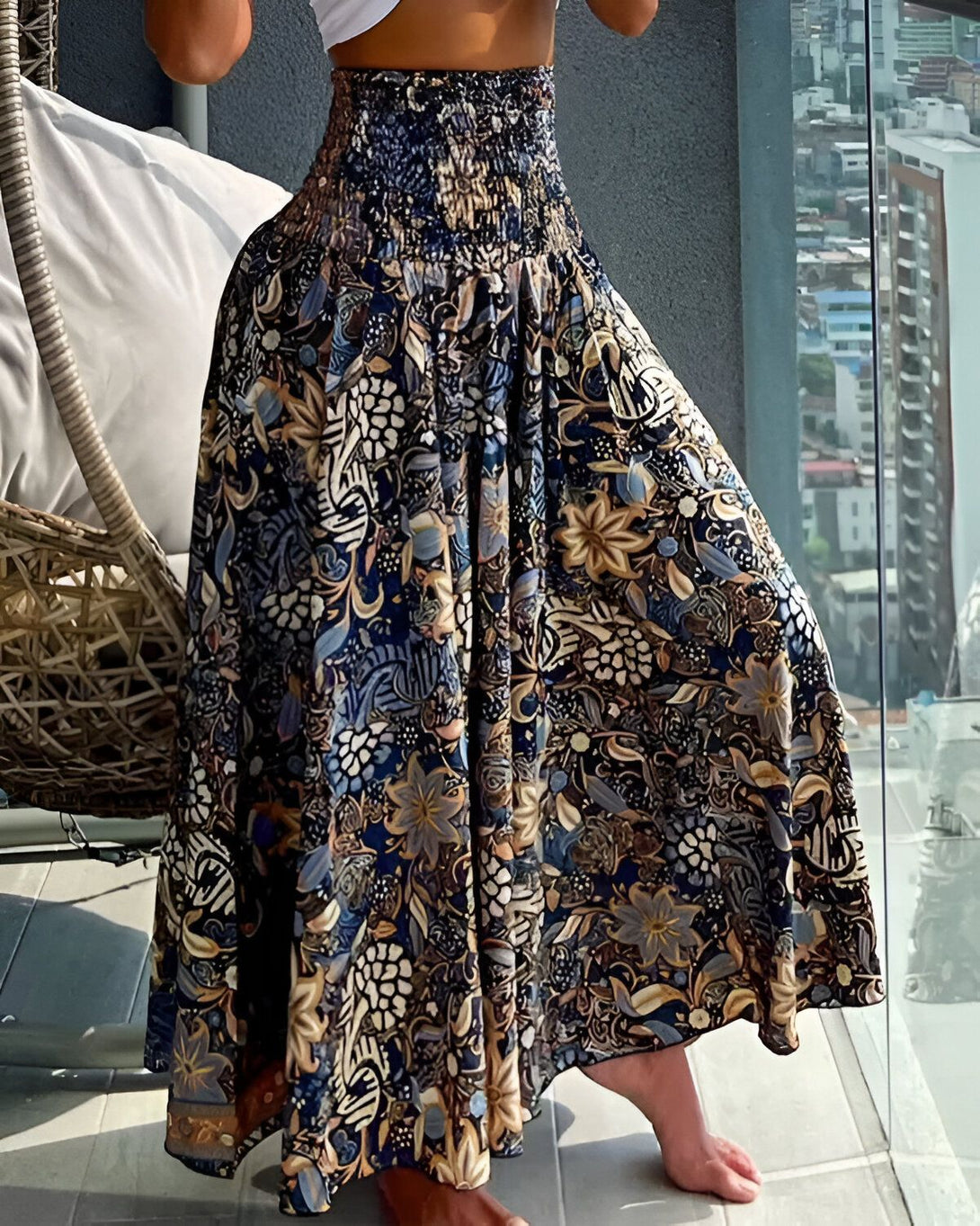 Une femme porte une jupe longue à taille haute. La jupe est bleu marine et marron, avec un motif floral. La femme est debout sur un balcon, avec une vue sur la ville en arrière-plan.