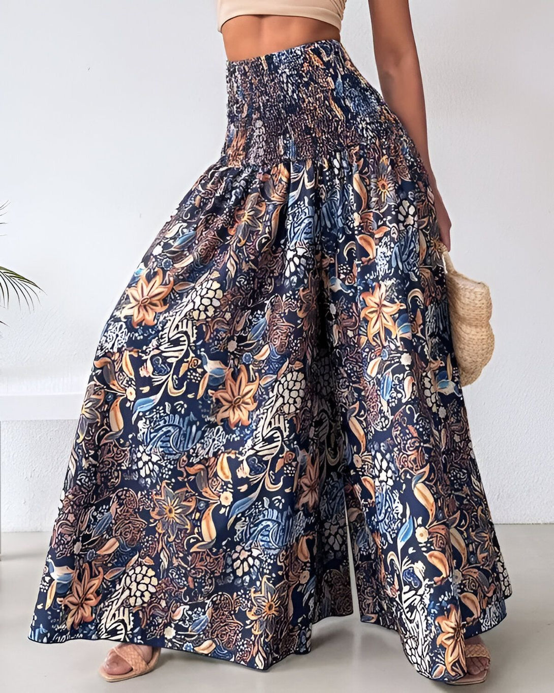 Une femme porte un pantalon large et coloré. Le pantalon est bleu marine et marron, avec un motif floral. Il est taille haute et a une ceinture élastiquée. La femme porte également un haut court beige et des sandales marron. Elle a les cheveux longs et bruns et porte un panier en osier à la main.