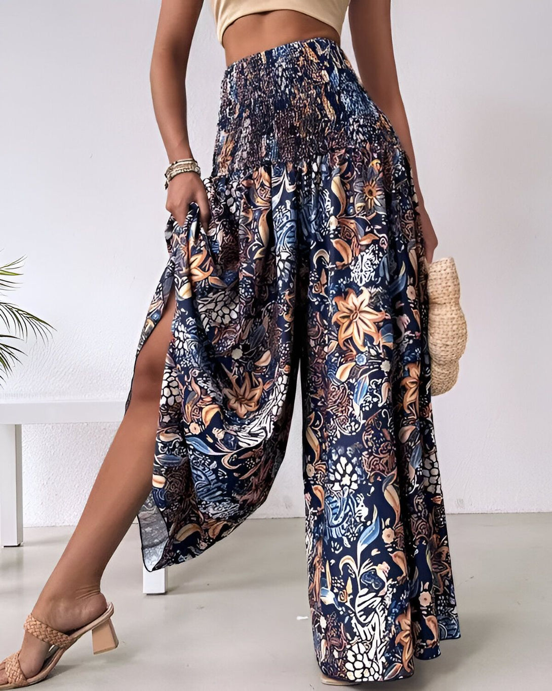 Une femme porte un pantalon large et coloré. Le pantalon est bleu marine et présente un motif floral coloré. La femme porte également un haut beige et des sandales marron. Elle a les cheveux longs et bruns et porte un sac à main en osier à la main droite.