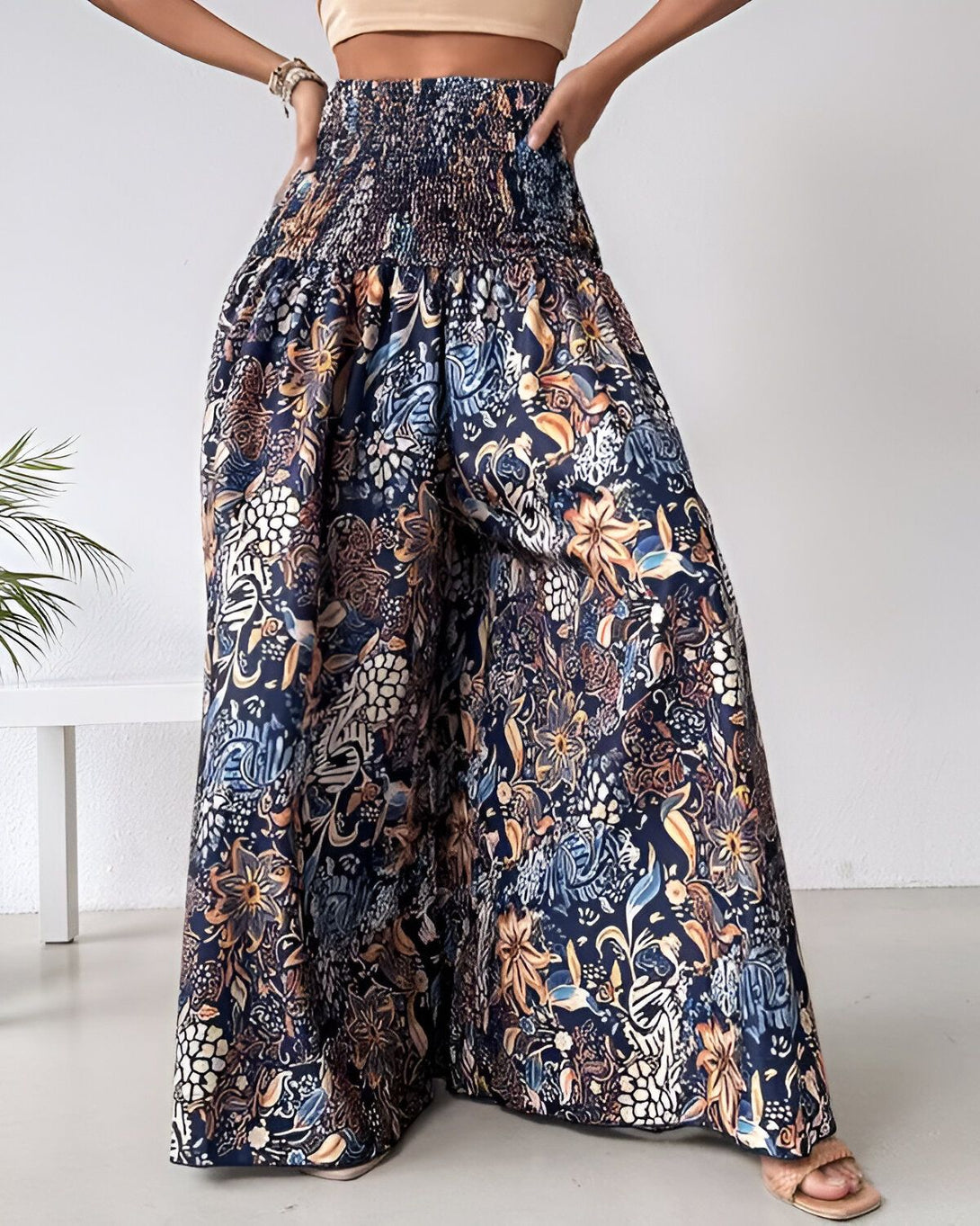 Une femme porte un pantalon large et coloré. Le pantalon est bleu marine et marron, avec des motifs floraux. La femme est vue de face et porte un haut beige. Elle a les cheveux longs et bruns.