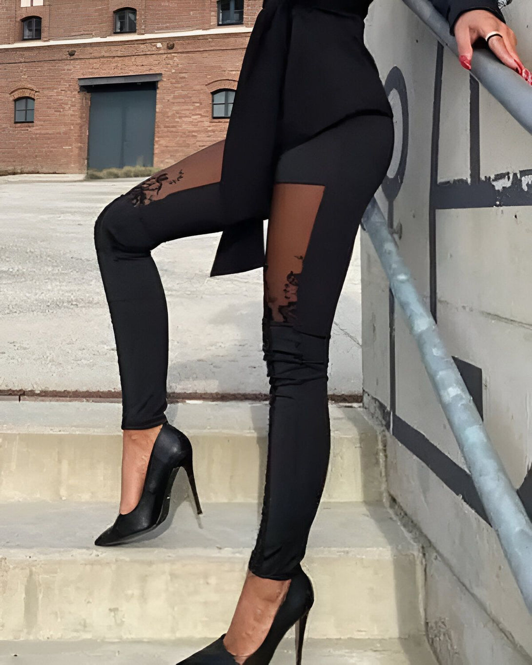 Une femme porte un legging noir avec des empiècements en dentelle sur les côtés. Elle porte également des escarpins noirs et une veste noire.