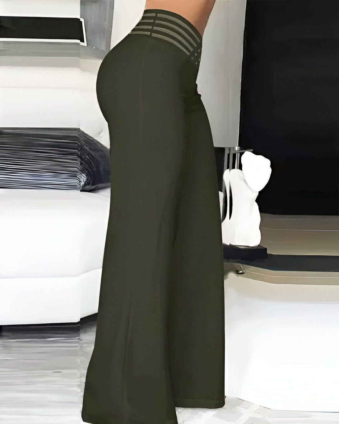 Une femme porte un pantalon taille haute vert olive. Le pantalon est évasé et a une ceinture à nouer. La femme est debout à côté d'un canapé blanc.