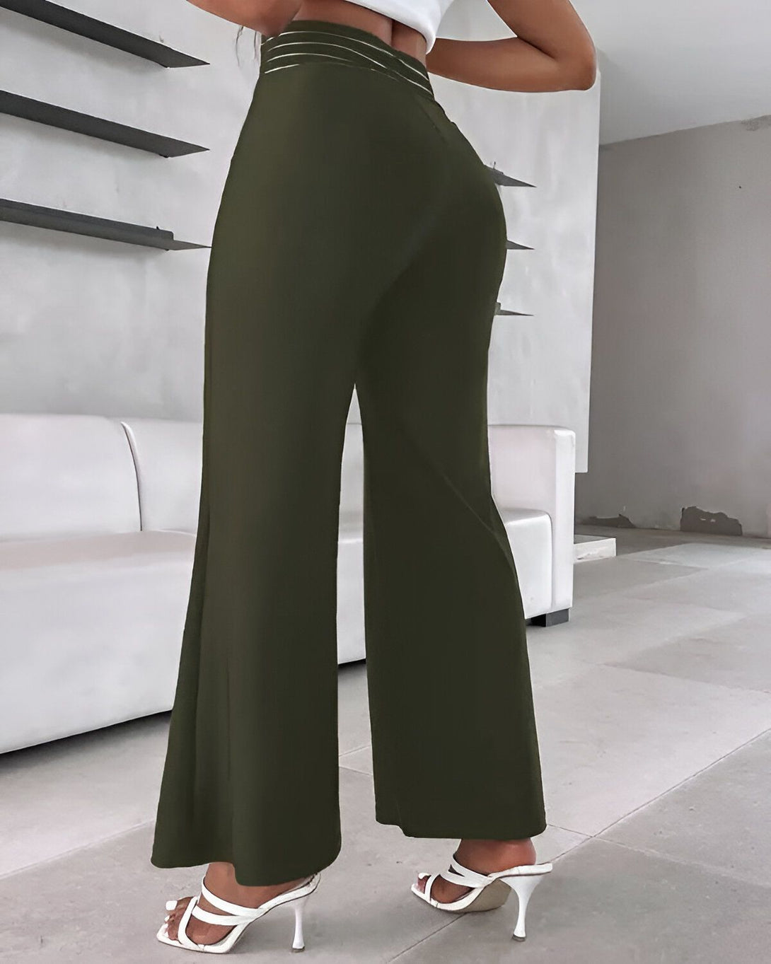 Une femme porte un pantalon vert olive taille haute et évasé. Elle est vue de dos et porte des sandales à talons blancs.