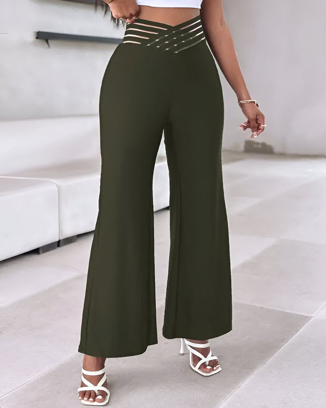 Une femme porte un pantalon taille haute vert olive avec une ceinture à lacets. Le pantalon est évasé et a une coupe ample. La femme porte également des sandales à talons blancs et un haut court blanc.