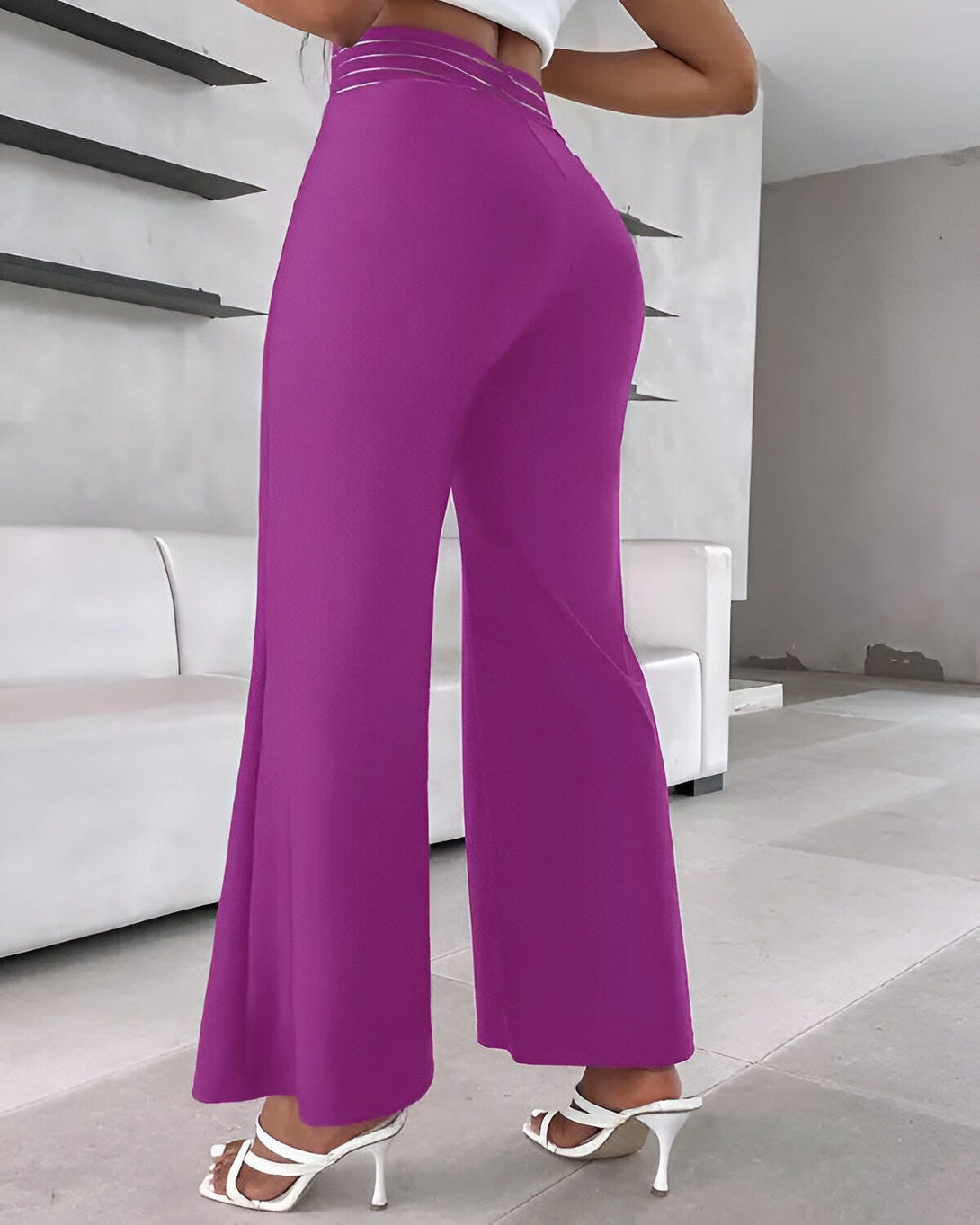 Une femme porte un pantalon large et évasé de couleur fuchsia. Le pantalon est taille haute et possède une ceinture. La femme est vue de dos et porte des sandales à talons blancs.