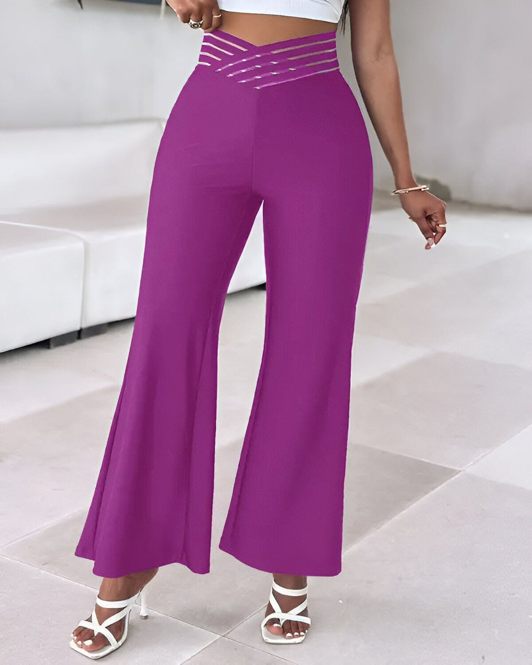 Une femme porte un pantalon taille haute évasé violet. Le pantalon est ajusté à la taille et évasé au niveau des hanches. Il est porté avec des talons blancs.