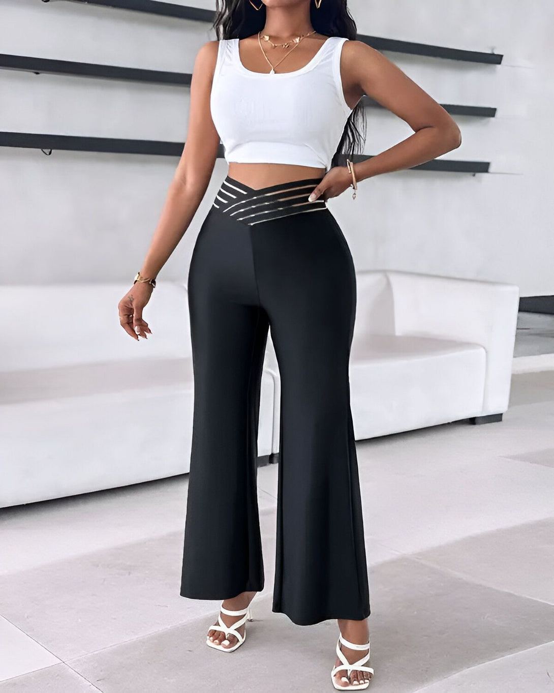 Une femme porte un pantalon taille haute noir avec une ceinture dorée et un haut blanc court. Elle porte également des sandales à talons blanches.