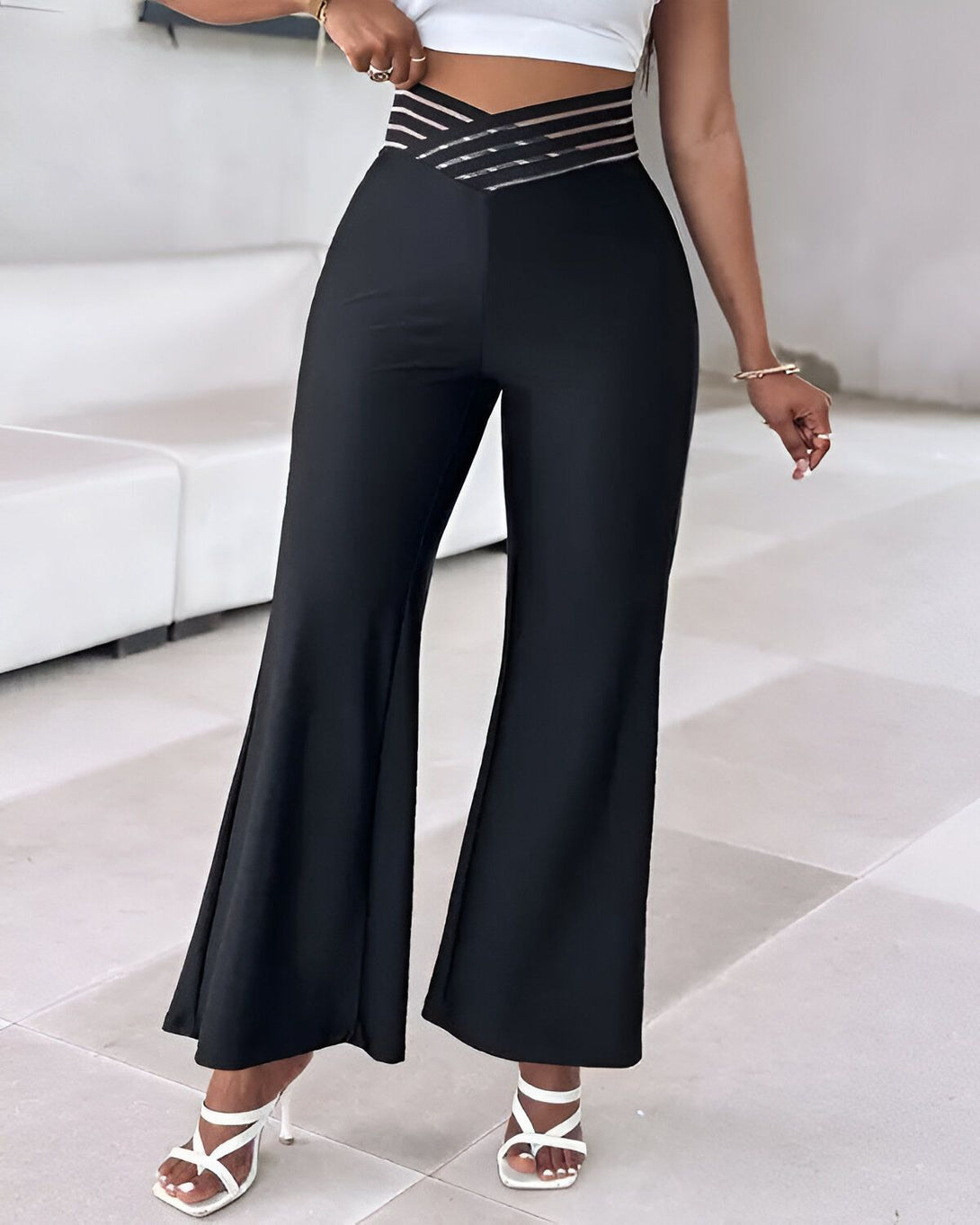 Une femme porte un pantalon noir taille haute évasé avec une ceinture transparente. Elle porte également un haut blanc et des sandales blanches.