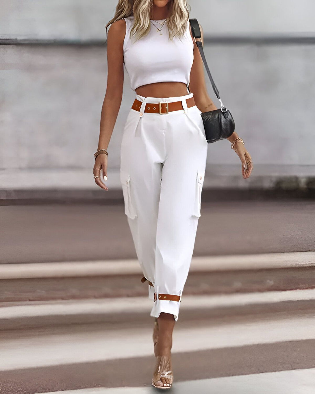 Une femme porte un pantalon cargo blanc taille haute avec une ceinture marron et un haut court blanc. Elle porte également des sandales à talons transparentes et un sac à main noir.