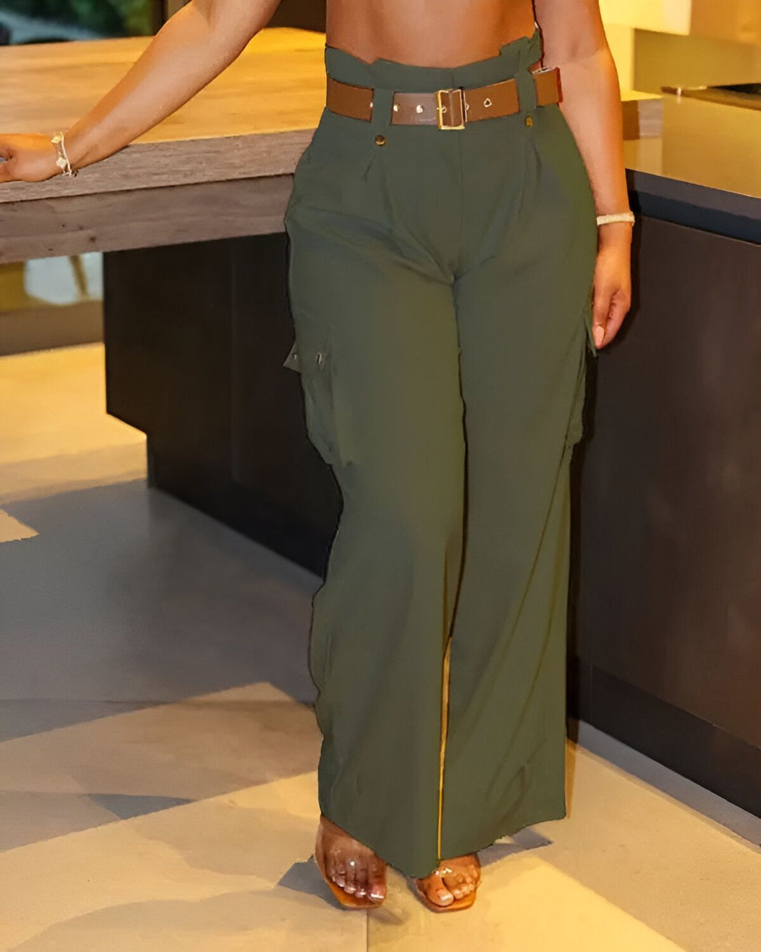 Une femme porte un pantalon cargo vert olive taille haute avec une ceinture marron. Elle est debout dans une cuisine et s'appuie sur un comptoir.